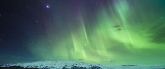 Aurora Borealis lights up the night sky with vibrant green and purple hues. Mountains, dusted with snow, stretch across the horizon beneath the dynamic display. Stars twinkle faintly in the dark sky above.