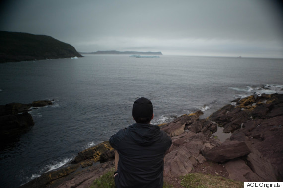 A person wearing a dark jacket and hat sits on rocky terrain, gazing out at a vast, cloudy seascape. The ocean is calm, with distant landforms visible under an overcast sky.